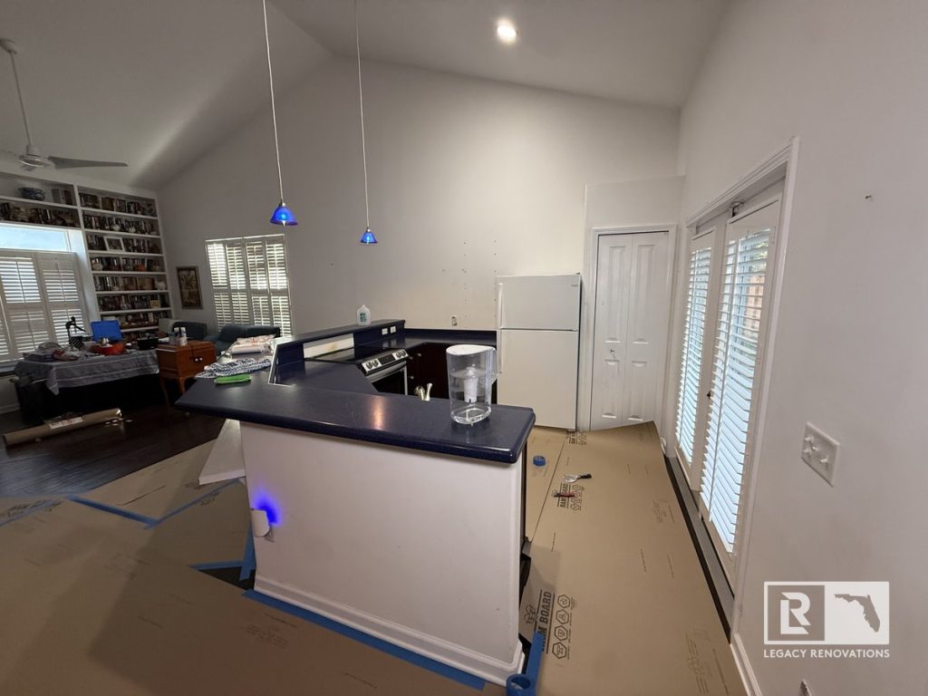 Open kitchen before renovation with a central peninsula, vaulted ceiling, pendant lights, patio doors, white refrigerator, and connected living area in a Jacksonville, Florida home.