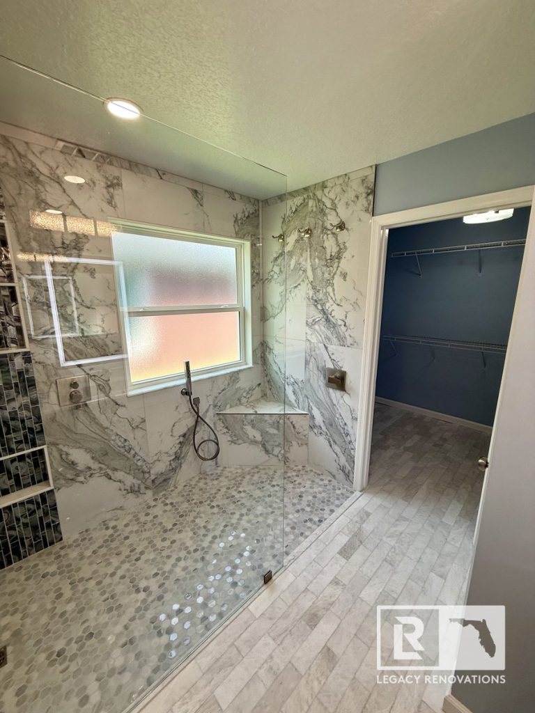 High-angle view of a luxury walk-in shower with marble-look porcelain tile and frameless glass next to an adjoining walk-in closet in a Jacksonville, FL bathroom.