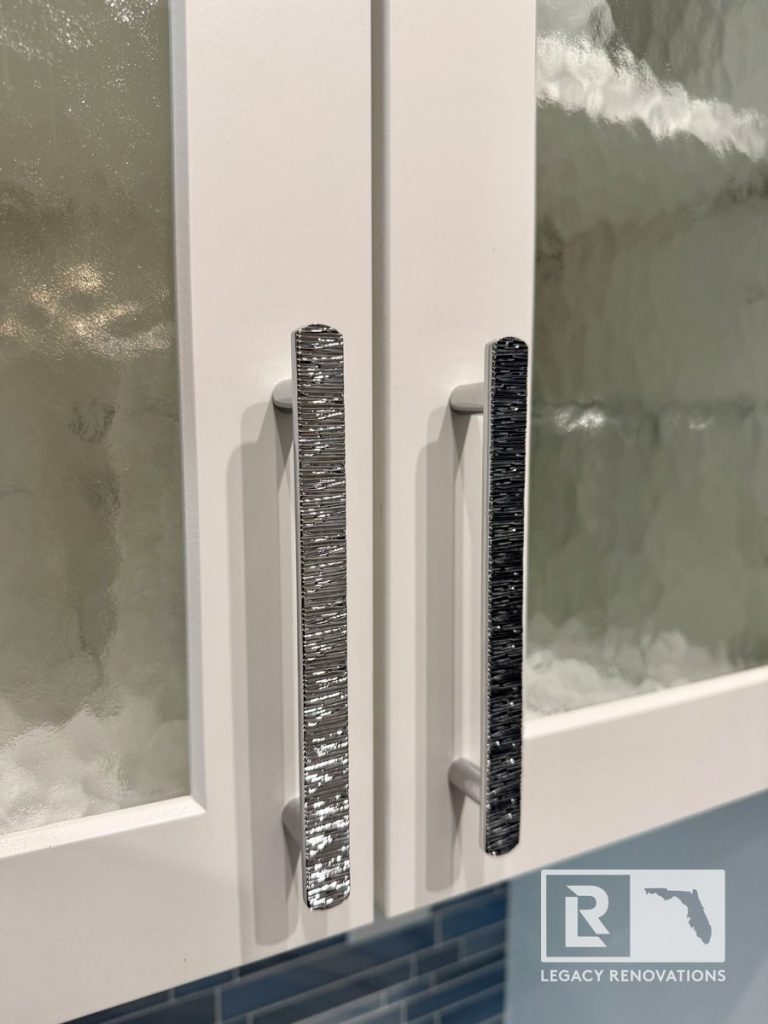 Close-up of white shaker kitchen cabinets with textured glass inserts, decorative brushed nickel cabinet handles, and a blue glass subway tile backsplash in a Jacksonville, FL kitchen remodel.
