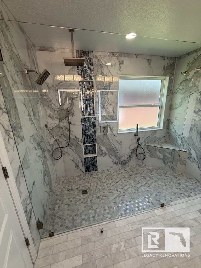 Wide-angle view of a large walk-in shower featuring frameless glass, double showerheads, rain head, marble wall tiles, and blue accent niche in Jacksonville, FL.