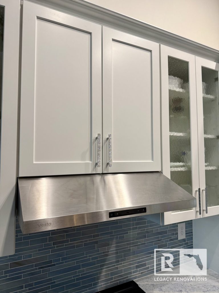 Kitchen remodel with white shaker upper cabinets, frosted glass cabinet doors, stainless steel range hood, blue glass subway tile backsplash, and brushed metal hardware in Jacksonville, FL.