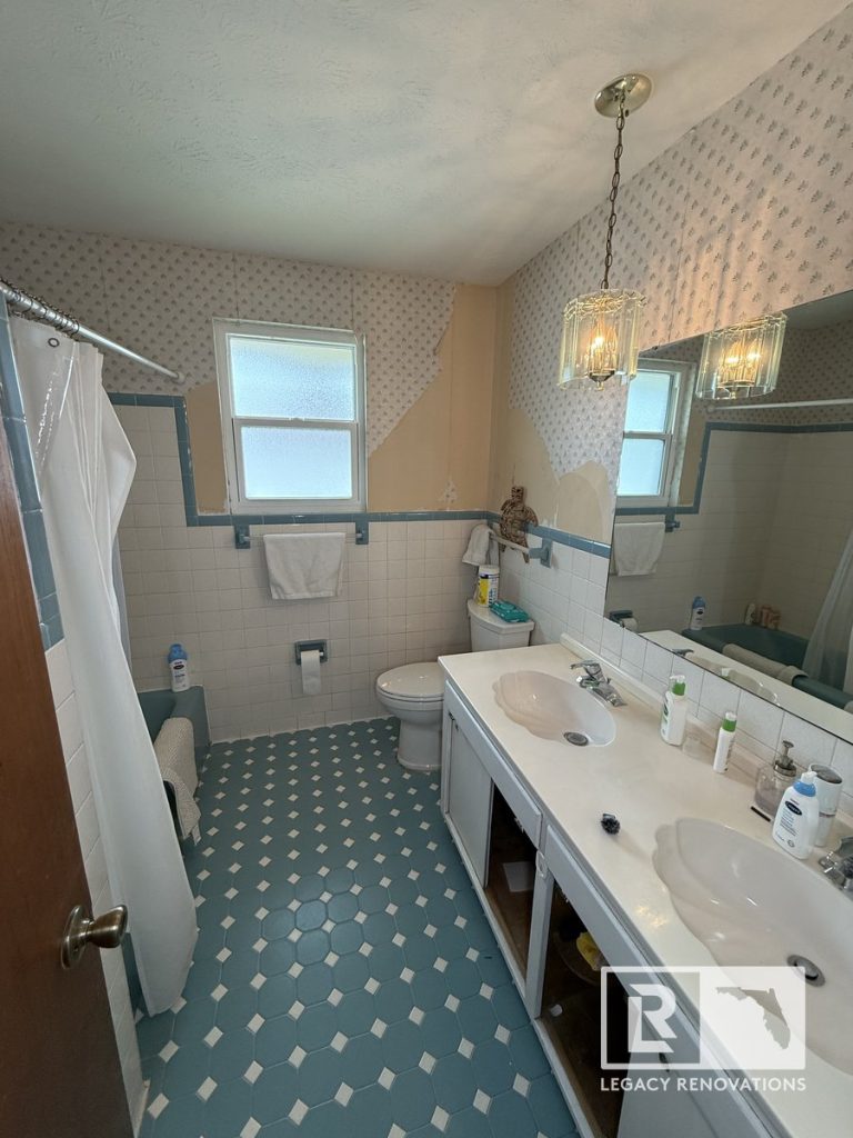 Before photo of a bathroom remodel in Jacksonville, Florida featuring an original double vanity, blue geometric floor tile, patterned wallpaper, and a bathtub with a shower curtain.