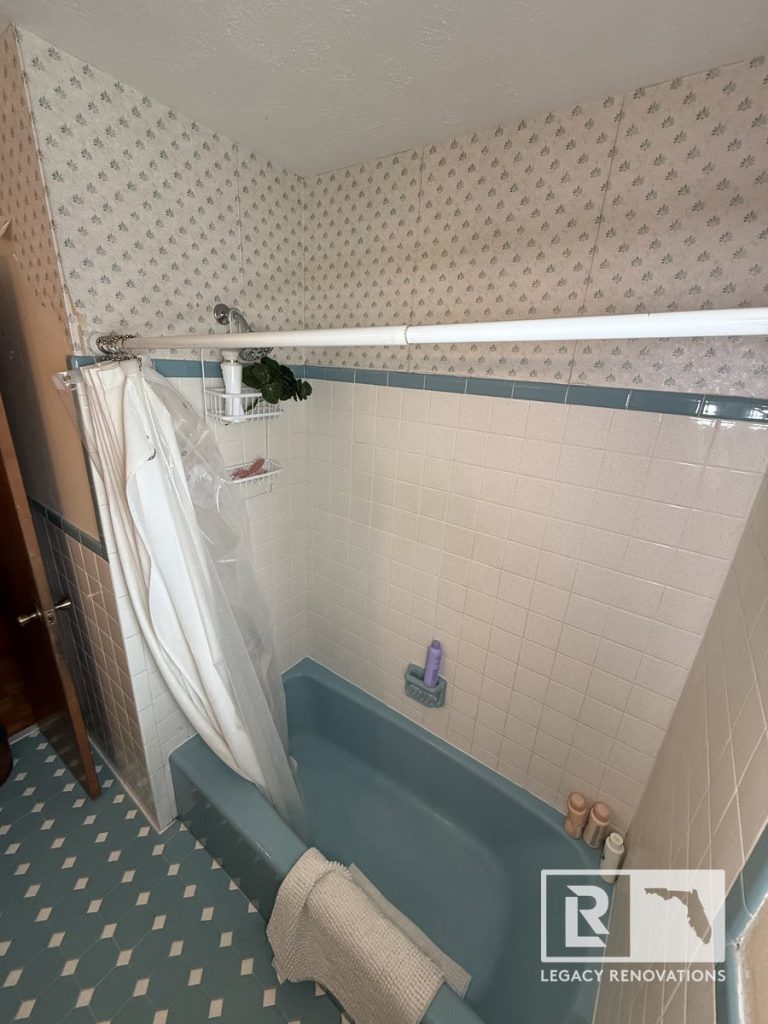 Before bathroom remodel in Jacksonville, Florida featuring an original blue bathtub, ceramic wall tile surround, patterned wallpaper, and vintage flooring.