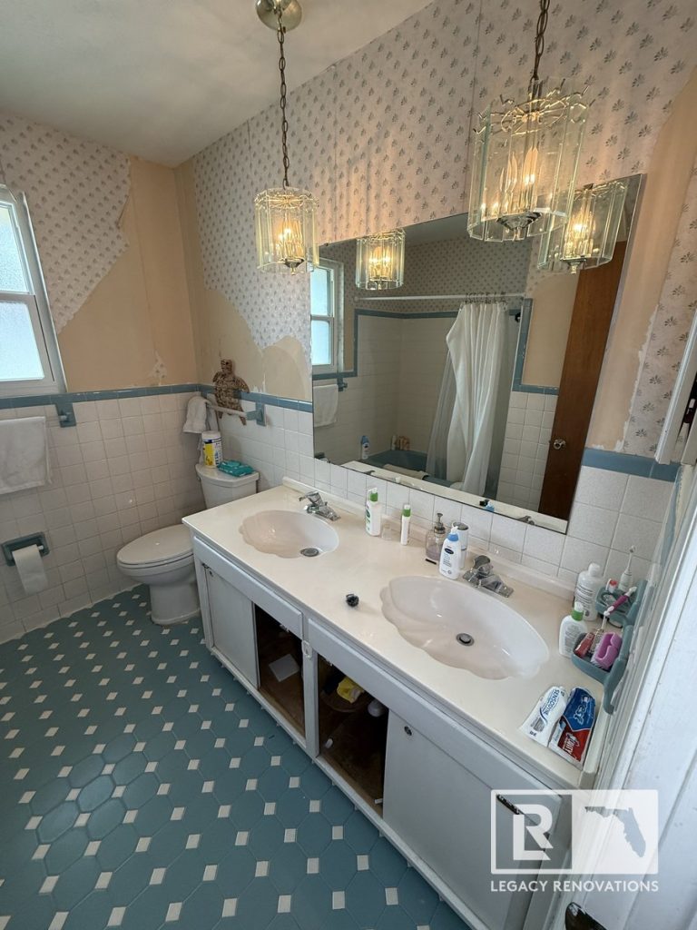 Before photo of a bathroom remodel in Jacksonville, Florida featuring an original double vanity, large wall mirror, patterned wallpaper, and blue geometric floor tile.