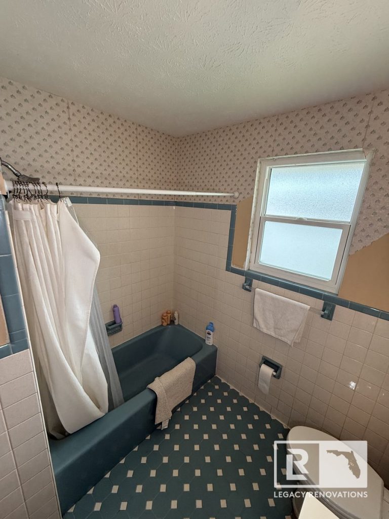 Original vintage bathroom in Jacksonville, Florida featuring a blue bathtub, retro wall tile, patterned wallpaper, frosted window, and classic flooring before remodeling.