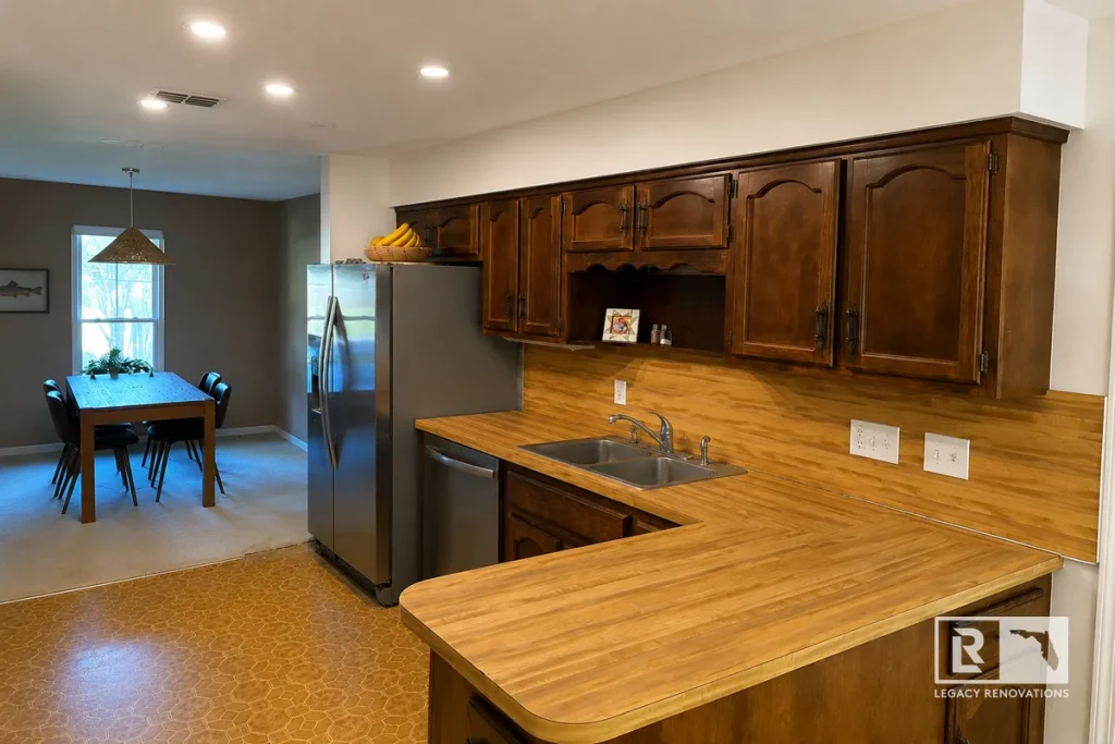 Wide angle view of an outdated kitchen with dark wood raised-panel cabinets, wood-grain laminate countertops, and patterned vinyl flooring before a remodel in Jacksonville, FL.