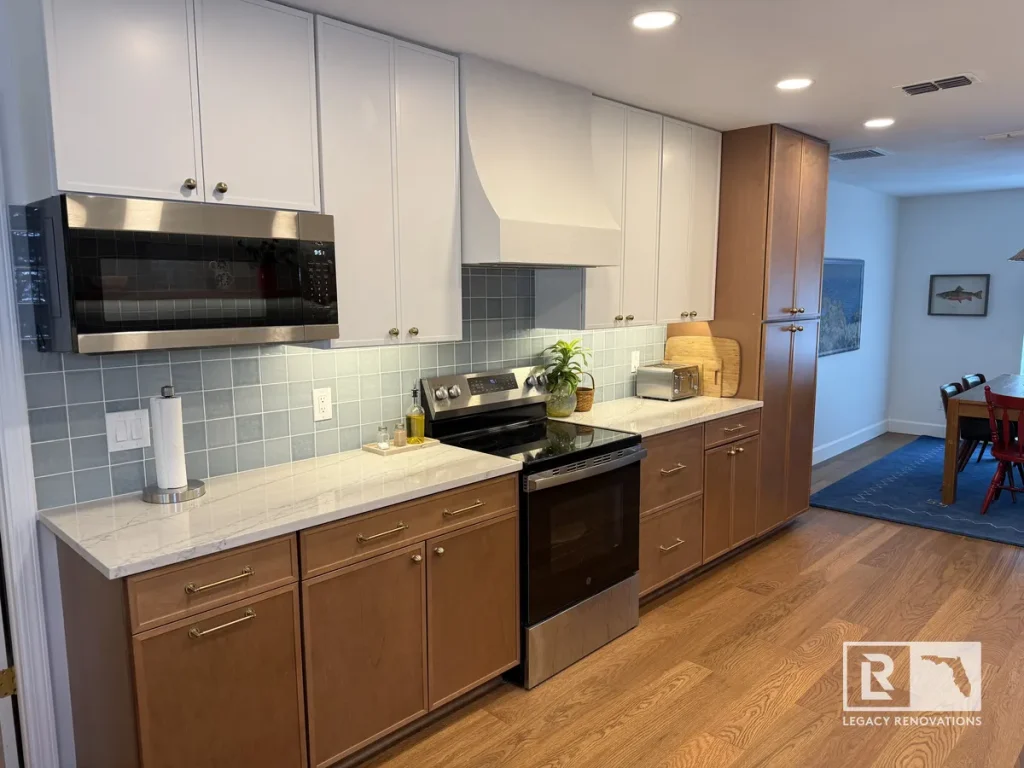 Full view of a remodeled kitchen featuring white upper cabinets, wood lower cabinets, quartz countertops, custom plaster hood, floating shelves, and blue tile backsplash.