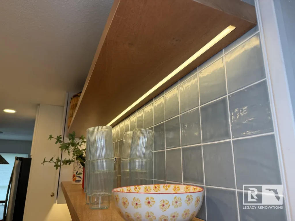 Floating wooden kitchen shelf with built-in LED strip light above a light blue ceramic tile backsplash.