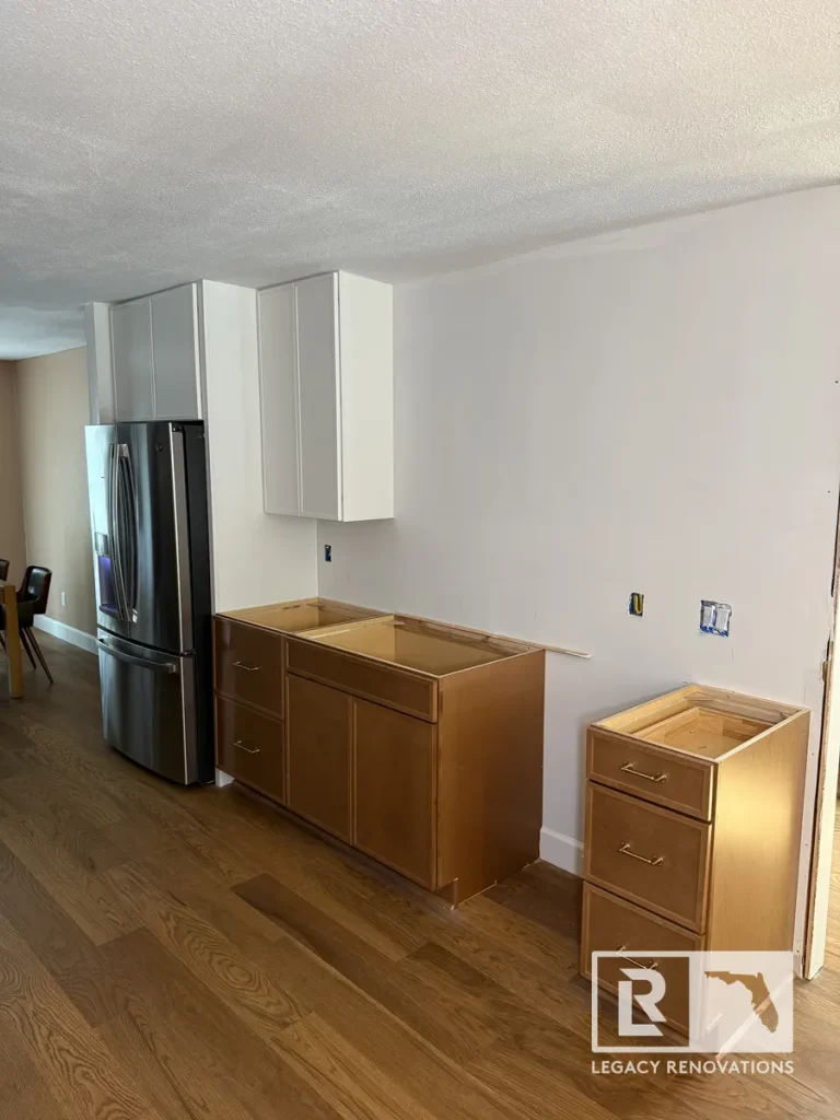 Mid-renovation kitchen in Jacksonville, FL showing installed white upper cabinets, natural wood base cabinets without countertops, and a new stainless refrigerator over wood floors.