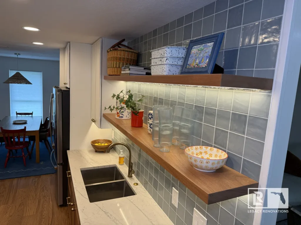 Modern kitchen featuring walnut floating shelves, handmade blue tile backsplash, quartz countertops, workstation sink, and warm under-cabinet lighting.