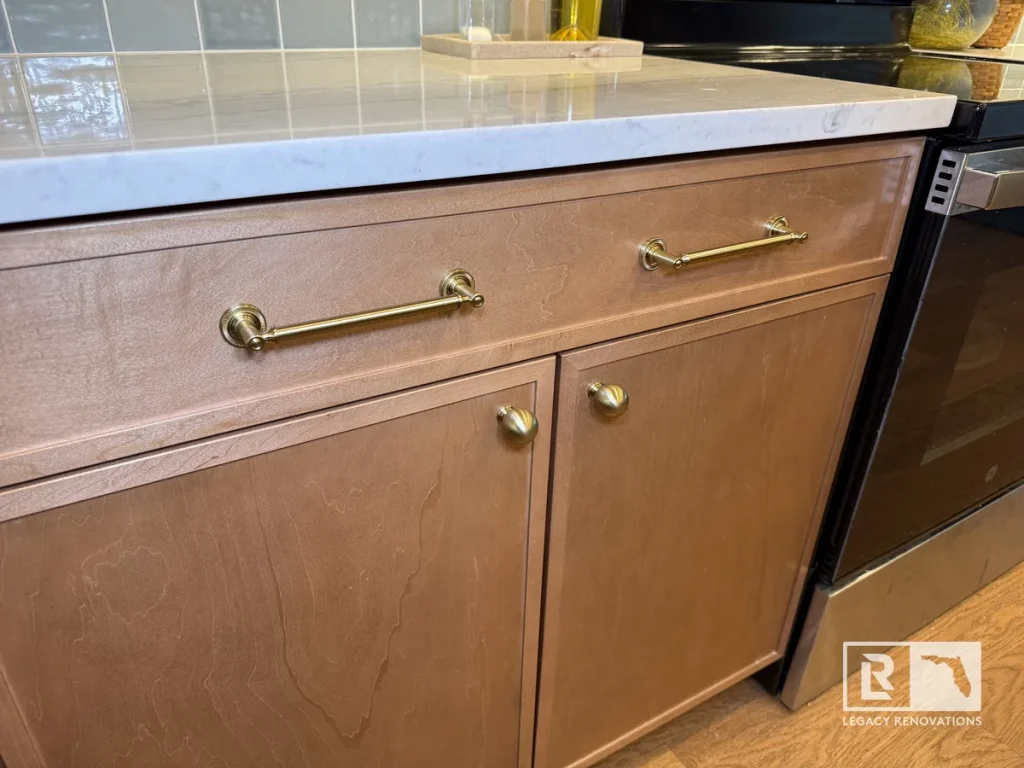 Close-up of custom light wood kitchen base cabinets featuring brushed brass pulls and knobs under a white marble countertop in Jacksonville, FL.