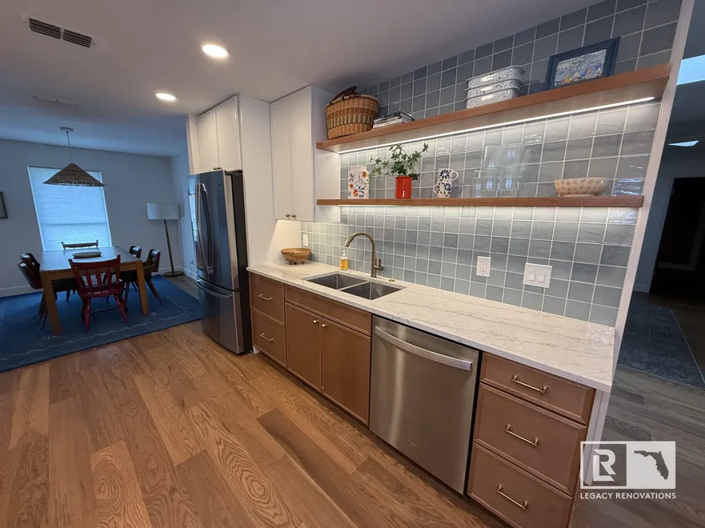 Wide-angle view of a modern kitchen remodel in Jacksonville, FL featuring natural wood base cabinets, stainless steel dishwasher, blue tile wall, and warm floating shelves.
