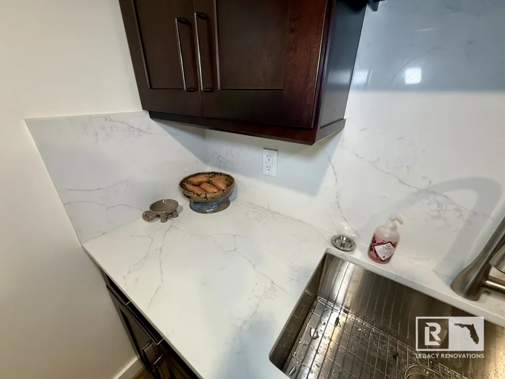 White quartz kitchen countertop and full-height backsplash with dark wood cabinets, stainless steel undermount sink, and modern faucet in a remodeled kitchen in Jacksonville Beach, FL.