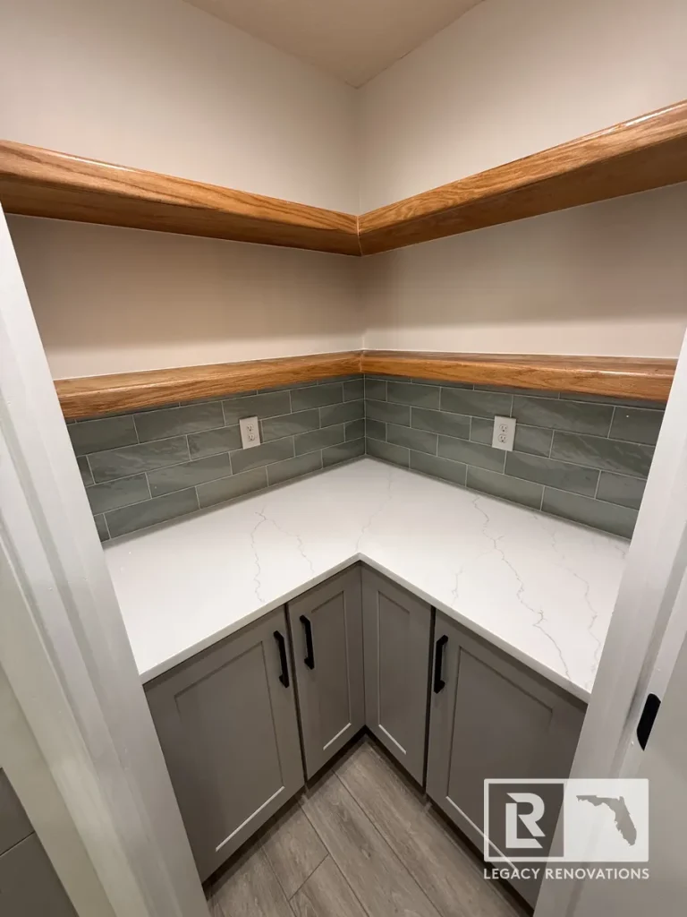 Corner view of gray cabinetry, white quartz countertop, sage subway tile backsplash, and floating wooden shelves in a Jacksonville FL home pantry.